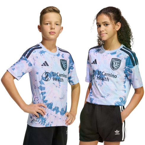 Two kids pose in matching light blue and pink tie-dye San Jose Earthquakes soccer jerseys and black shorts against a solid black background.