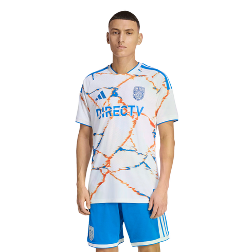 A man models a San Diego FC soccer kit with a white jersey featuring orange and blue abstract patterns and a DIRECTV logo, paired with blue shorts.