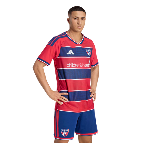 Male model posing in the FC Dallas home soccer kit, featuring a red and blue striped jersey with "children'shealth" logo and matching blue shorts.