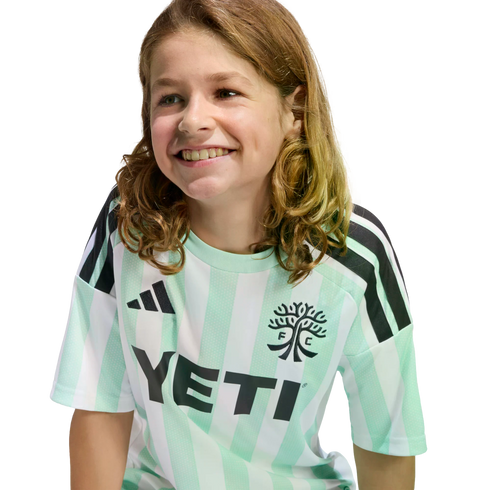 A smiling child with long, wavy brown hair wearing a mint green and white striped Austin FC jersey with a black Yeti logo, set against a solid black background.