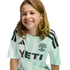 A smiling child with long, wavy brown hair wearing a mint green and white striped Austin FC jersey with a black Yeti logo, set against a solid black background.