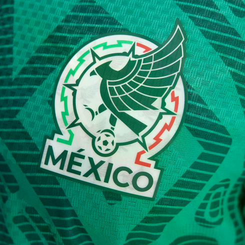 Close-up of the Mexico national football team crest on a green textured jersey. The emblem features a stylized eagle, a soccer ball, and the word "MÉXICO".