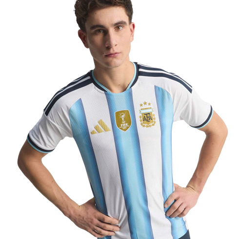 A man wearing the Argentina national football team home jersey with light blue and white stripes and gold accents, including the 2022 FIFA World Champions patch, posing with hands on his hips.