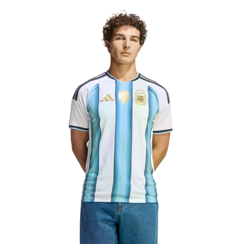 A man with curly hair wearing a blue and white striped Argentina national football team jersey and jeans, standing against a black background.