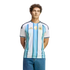 A man with curly hair wearing a blue and white striped Argentina national football team jersey and jeans, standing against a black background.