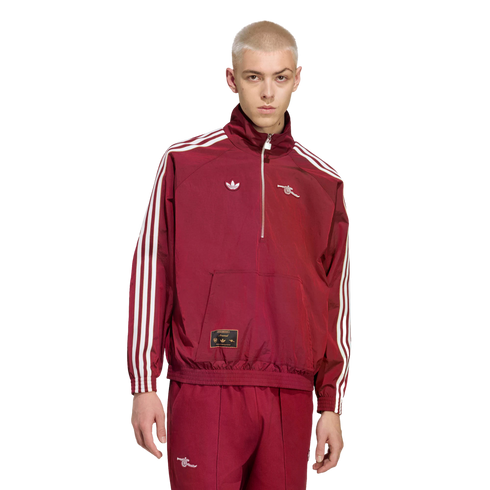 A person with short blonde hair models a maroon Adidas x Arsenal tracksuit with white stripes and logos against a solid black background.