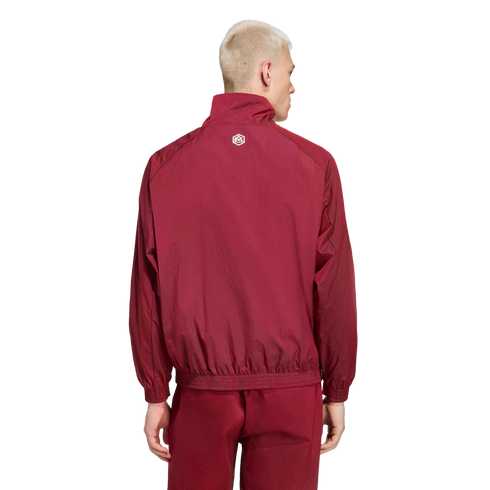 Back view of a man with short blond hair wearing a maroon tracksuit against a black background. The jacket features a small white logo on the upper back.