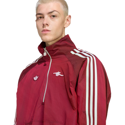 A blond man wears a maroon Adidas x Arsenal half-zip track jacket with white stripes on the sleeves, set against a black background.