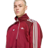 A blond man wears a maroon Adidas x Arsenal half-zip track jacket with white stripes on the sleeves, set against a black background.