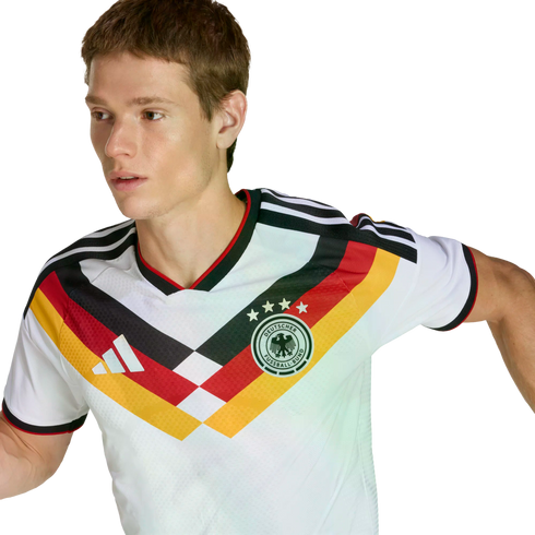 A man wearing a white German football jersey featuring a black, red, and yellow chevron design, looking to his left against a solid black background.