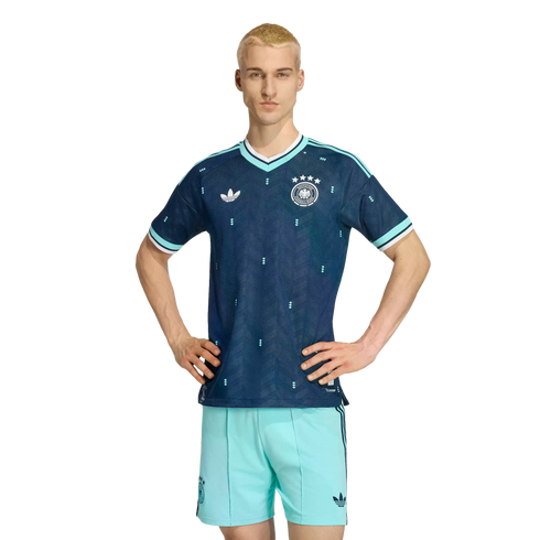 A man models a navy blue and teal German national team soccer kit, standing with hands on his hips against a solid black background.