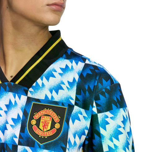 Close-up of a Manchester United jersey with a blue and white jagged geometric pattern and the club crest, featuring a black collar with yellow trim.