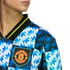 Close-up of a Manchester United jersey with a blue and white jagged geometric pattern and the club crest, featuring a black collar with yellow trim.