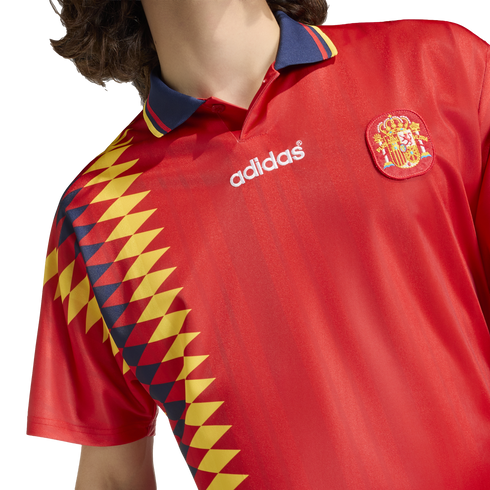 Red Spain Adidas football jersey with a blue polo collar, the national crest, and a vertical yellow and navy diamond pattern on the side.