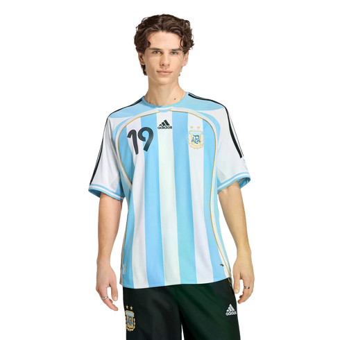 A young man with curly brown hair wearing a blue and white striped Argentina football jersey and black shorts against a black background.