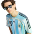 A young man with wavy hair and blue-tinted glasses wears an Argentina national team jersey featuring the number 19 against a solid black background.