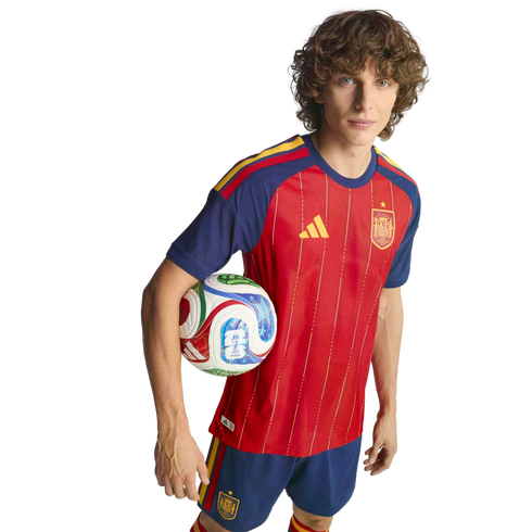 A young man in a red and blue Spain national football kit holds a soccer ball under his arm against a black background.