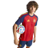 A young man in a red and blue Spain national football kit holds a soccer ball under his arm against a black background.