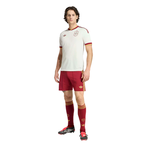 A man models a white and maroon Adidas Spain national football team kit against a black background.
