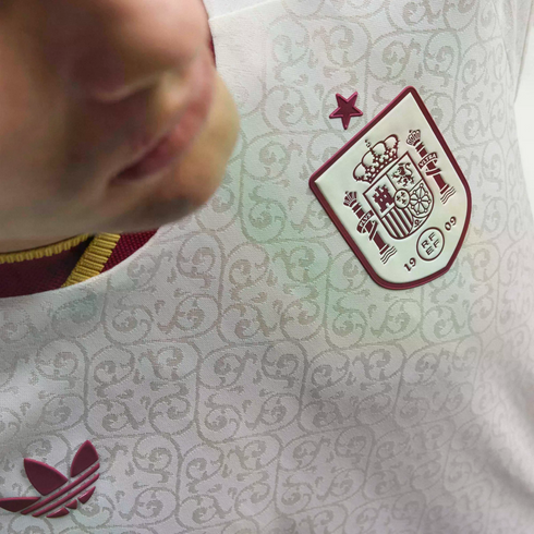 Close-up of a white Spain national team soccer jersey with a subtle ornate pattern, featuring a maroon RFEF crest and star, and an Adidas trefoil logo.