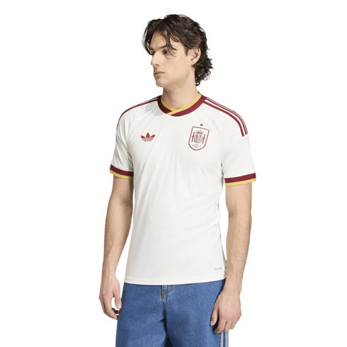 A man modeling a white Spain national football team jersey with red and yellow trim, paired with blue jeans, against a solid black background.