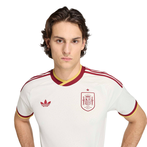 Young man with wavy brown hair in a white Spain national football team jersey with burgundy and yellow trim, against a black background.