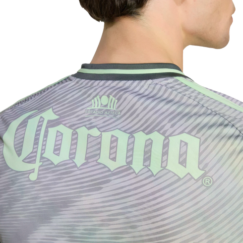 A close-up of the back of a person wearing a light green and grey patterned sports jersey with an Estadio Azteca emblem and a large Corona logo.