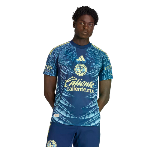 A Black man wearing a blue and yellow Club América soccer kit featuring intricate feather patterns and sponsor logos, posed against a plain black background.