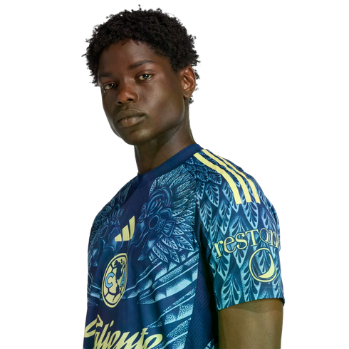 A Black man with short curly hair wears a patterned blue and yellow soccer jersey, looking at the camera against a black background.
