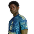 A Black man with short curly hair wears a patterned blue and yellow soccer jersey, looking at the camera against a black background.