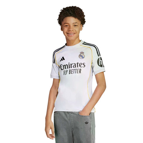 Smiling young person with curly hair and braces, wearing a white Real Madrid jersey and grey pants, isolated on a white background.