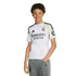 Smiling young person with curly hair and braces, wearing a white Real Madrid jersey and grey pants, isolated on a white background.
