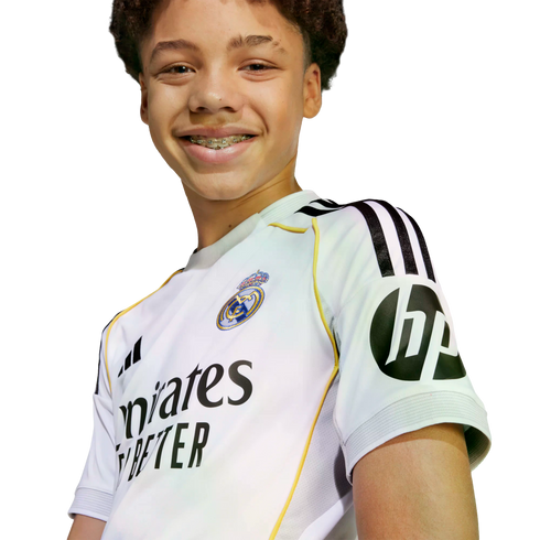 A smiling young boy with curly hair and braces wearing a white Real Madrid soccer jersey against a plain background.