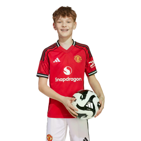 A smiling young boy with braces wearing a red Manchester United soccer jersey and white shorts while holding a soccer ball against a white background.