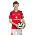 A smiling young boy with braces wearing a red Manchester United soccer jersey and white shorts while holding a soccer ball against a white background.