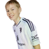 A young person with blonde hair smiles while leaning forward, wearing a white soccer jersey featuring several logos. The image is set against a solid black background.