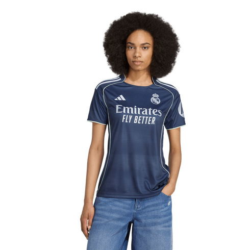 A woman with a large afro wears a navy blue Real Madrid soccer jersey and blue jeans against a white background.