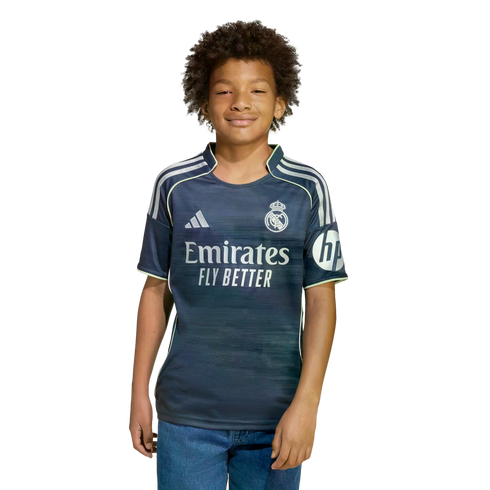 A boy with dark curly hair wearing a navy blue Real Madrid soccer jersey and blue jeans, smiling against a plain white background.