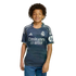 A boy with dark curly hair wearing a navy blue Real Madrid soccer jersey and blue jeans, smiling against a plain white background.