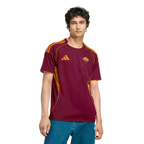 A man wearing a burgundy and orange AS Roma football jersey and blue jeans against a black background.