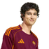 A young man with dark curly hair wearing a maroon and orange AS Roma soccer jersey, smiling against a black background.