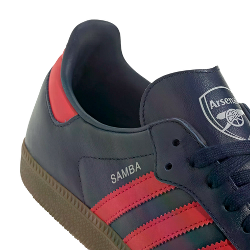 Close-up of a navy blue Adidas Samba sneaker featuring pink stripes and the Arsenal Football Club crest on the tongue.