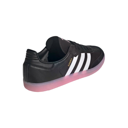 Rear-three-quarter view of an Adidas Samba sneaker featuring a black leather upper with three white stripes and a translucent pink gum sole.