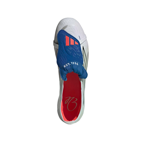 Top-down view of a white Adidas soccer cleat featuring a blue fold-over tongue with red stripes and a red insole with a cursive signature.