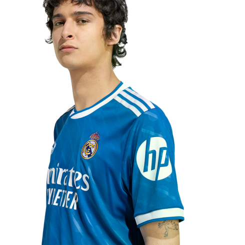 Young man wearing a blue Real Madrid soccer jersey with white trim and sponsor logos, posed against a black background.