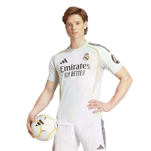 A man in a white Real Madrid kit poses with a soccer ball against a white background, holding the ball in one hand and resting the other on his hip.
