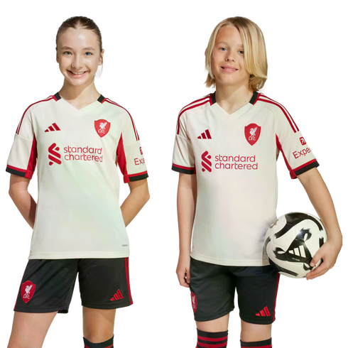 A girl and a boy wearing matching Liverpool FC jerseys and shorts smile against a black background. The boy holds a soccer ball.
