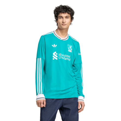 A man with dark curly hair wears a long-sleeved teal Adidas Liverpool FC jersey and dark blue jeans against a white background.