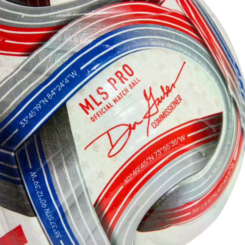 Close-up of a white MLS PRO soccer ball with red and blue stripes, the commissioner’s signature, and geographic coordinates printed on the surface.