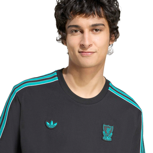 A man with dark wavy hair wearing a charcoal sweatshirt with teal shoulder stripes, a teal Adidas Trefoil logo, and a teal Liverpool FC crest on the chest.